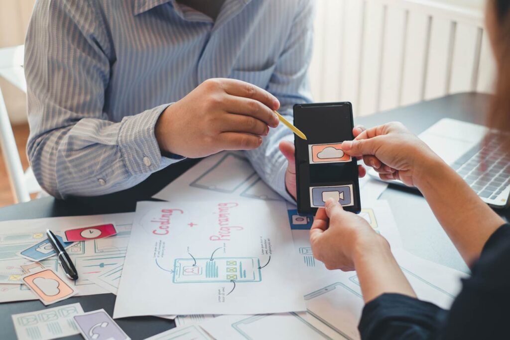 Two people collaborate on Android app mockups at a development agency in London, using a smartphone and design sketches.