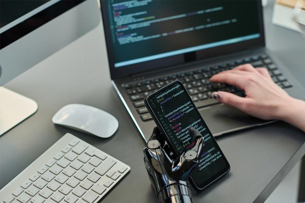Robotic hand holding smartphone with code displayed, next to a laptop and keyboard. Mobile Application Services in London.
