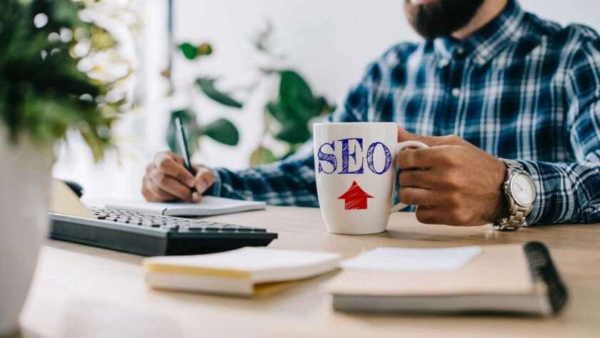 Bearded man writing, holding an "SEO" mug, representing the No.1 SEO Agency in London, at desk with laptop and notebook.